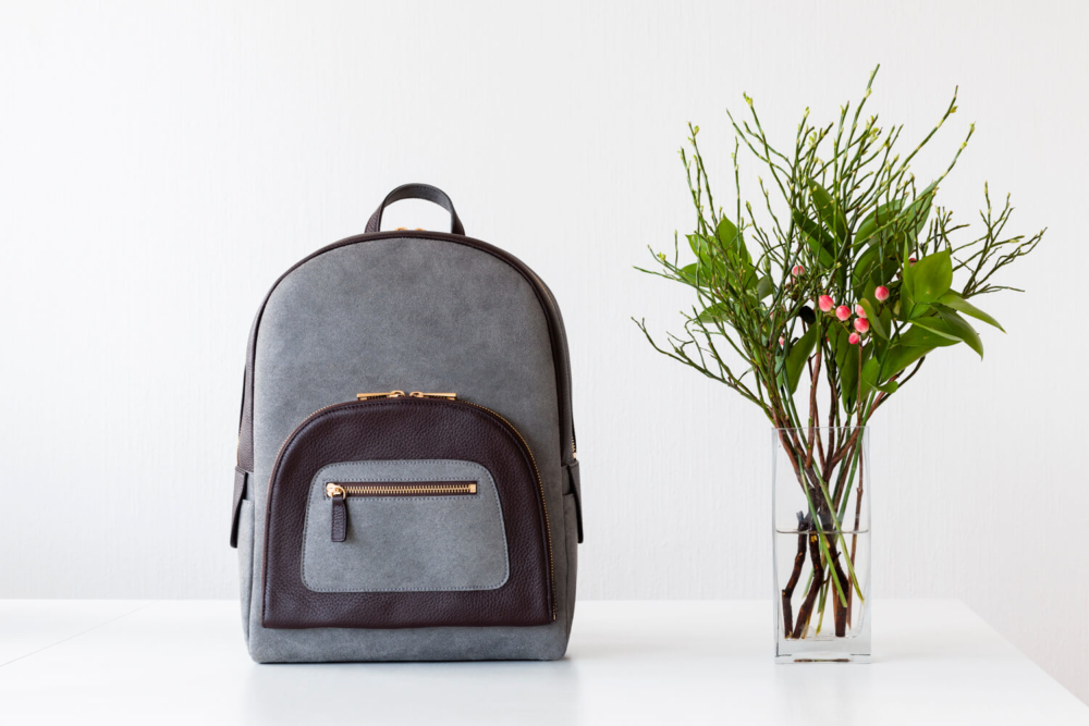 Un sac à dos gris et marron tendance se trouve sur une table blanche à côté d'un vase transparent rempli de branches vertes et de baies roses, le tout sur un fond blanc.