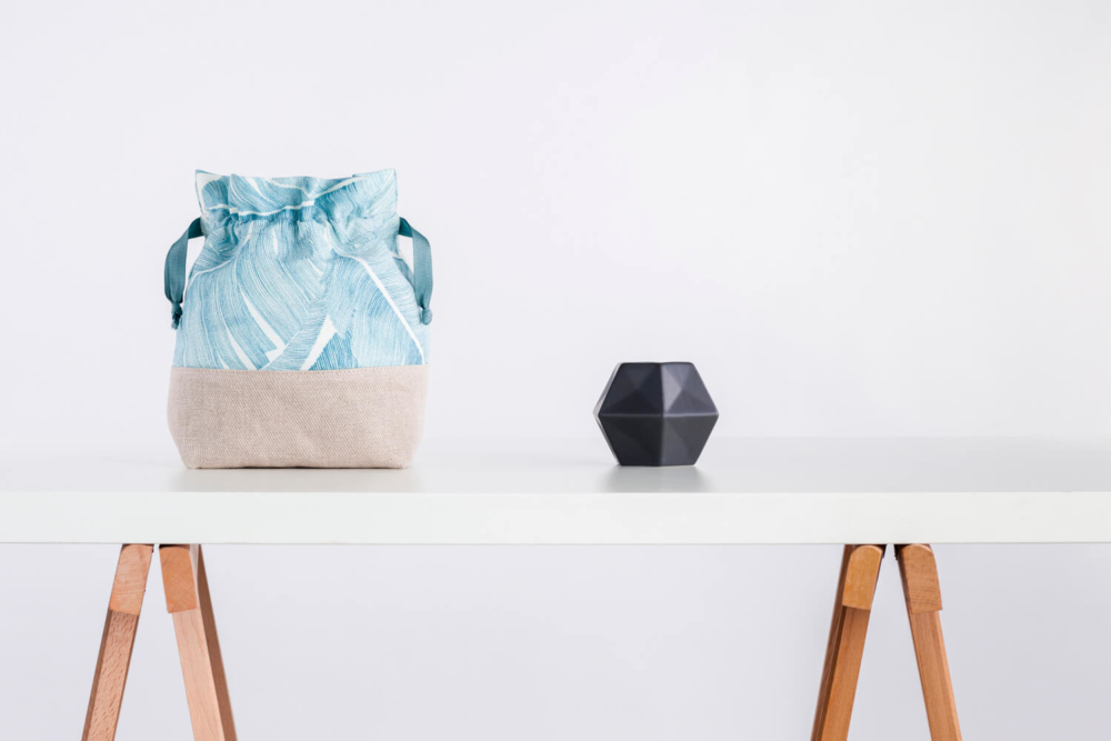 Un sac fourre-tout en tissu tendance avec un motif de feuilles bleues se trouve sur une table en bois moderne à côté d'un vase noir aux motifs géométriques. Le fond est blanc uni.