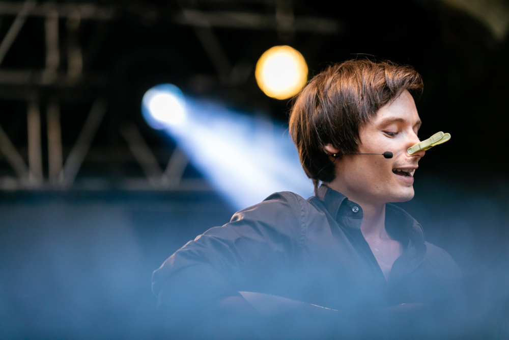 Un jeune homme chante avec passion sur scène avec un micro-casque. La scène a des lumières bleues vives et un projecteur brillant en arrière-plan.