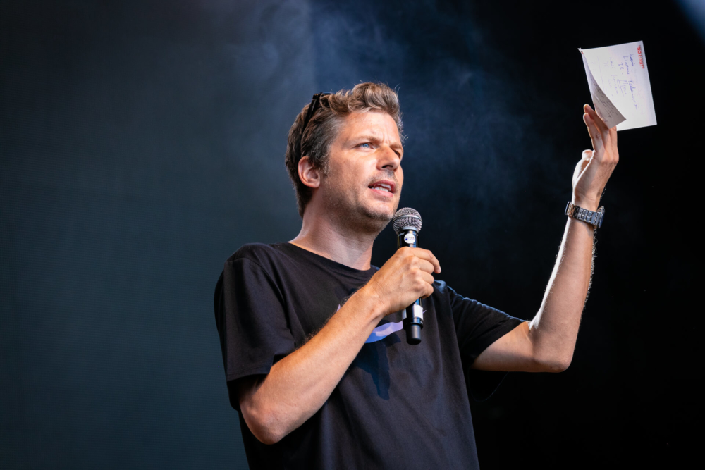 Un homme parle avec passion sur une scène extérieure, tenant un microphone et un morceau de papier, avec des effets de fumée derrière lui.