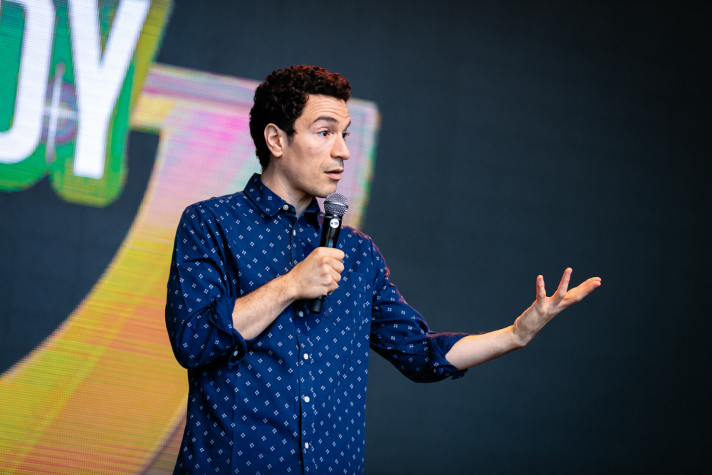 Un homme est debout sur une scène, parle et fait des gestes d’une main tout en tenant un microphone. Il porte une chemise bleue à motifs. L’arrière-plan est doté de lumières de scène lumineuses et d’écrans numériques.