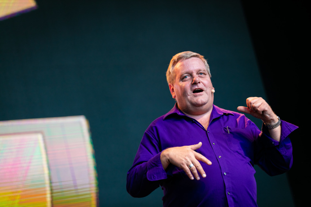 Un homme en chemise violette parle avec enthousiasme sur scène, utilisant ses mains pour faire des gestes. Derrière lui, un grand écran diffuse des images colorées.
