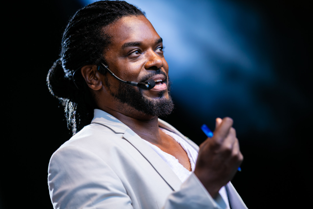 Un conférencier motivateur avec des dreadlocks sourit lorsqu'il prend la parole lors d'une conférence. Il porte un casque micro, une chemise blanche et une veste, sous les lumières bleues de la scène.