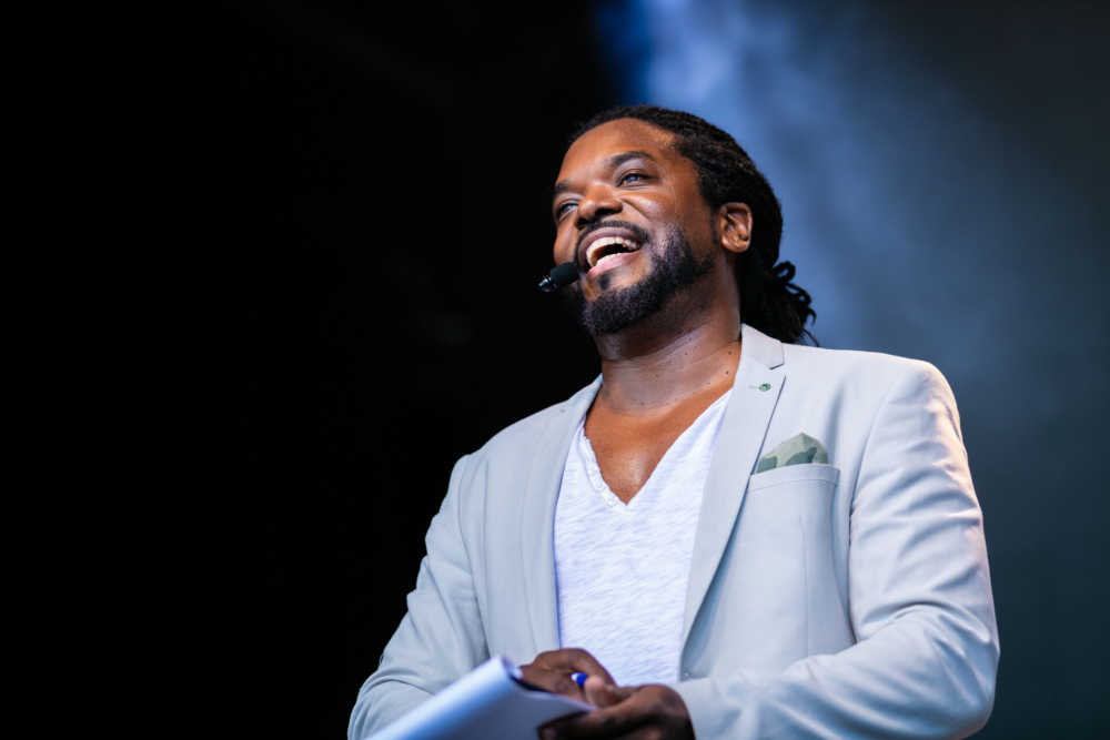 Un charmant homme en costume blanc et t-shirt parle dans un micro-casque. Il sourit largement sous les lumières vives de la scène sur un fond légèrement enfumé.