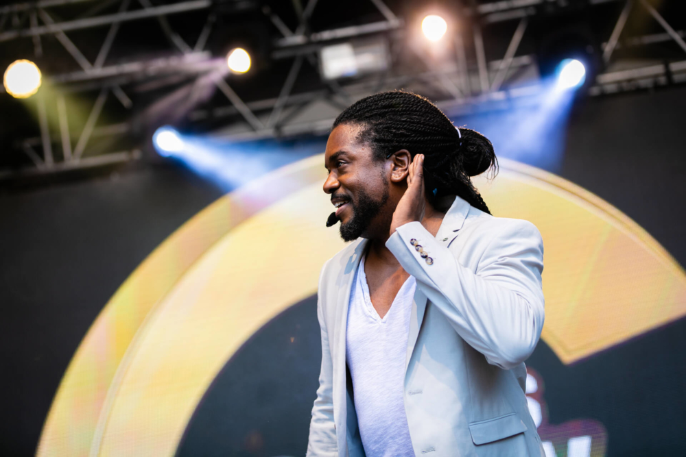 Un gars aux cheveux tressés, vêtu d'un costume gris et d'une chemise blanche, sourit et se touche l'oreille alors qu'il se produit sur une scène bien éclairée.