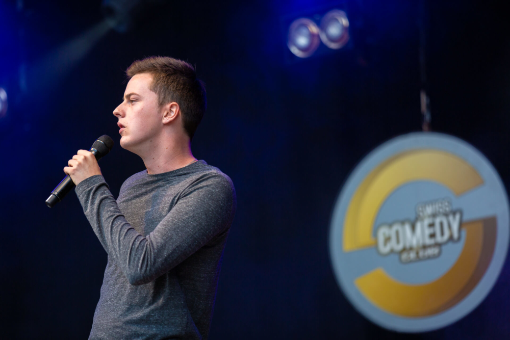 Un homme est sur scène lors d’un spectacle humoristique. Il tient un microphone et il y a un logo 