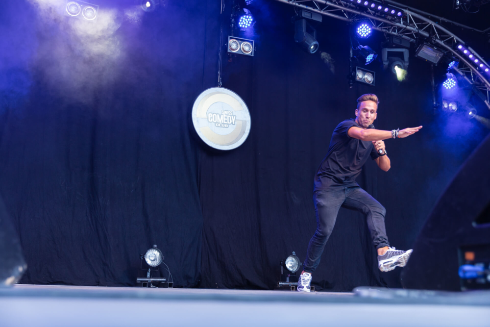 Un artiste d’un club de comédie est sur scène, agitant dramatiquement les deux bras et se tenant en équilibre sur une jambe. Les lumières et l'équipement bleus de la scène sont en arrière-plan.