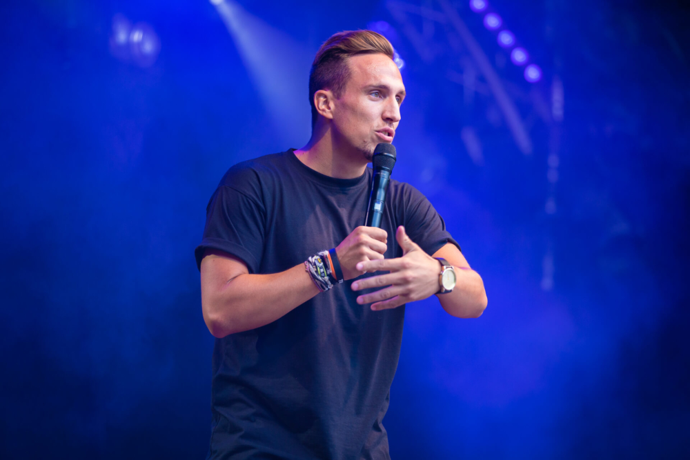 Un homme se tient sur une scène avec des lumières bleues et des effets de concert, parlant dans un microphone. Il porte un t-shirt noir et une montre et bouge sa main gauche pendant qu'il parle.