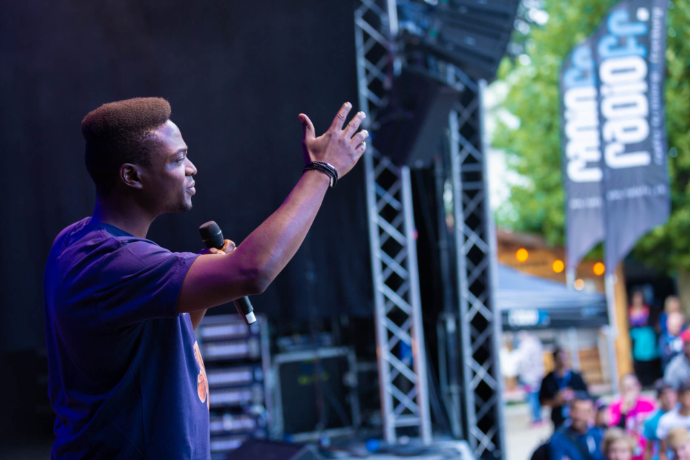 Un homme s’adresse avec enthousiasme à une foule lors d’un événement en plein air depuis une scène, à l’aide d’un microphone. Le public est visible au premier plan.