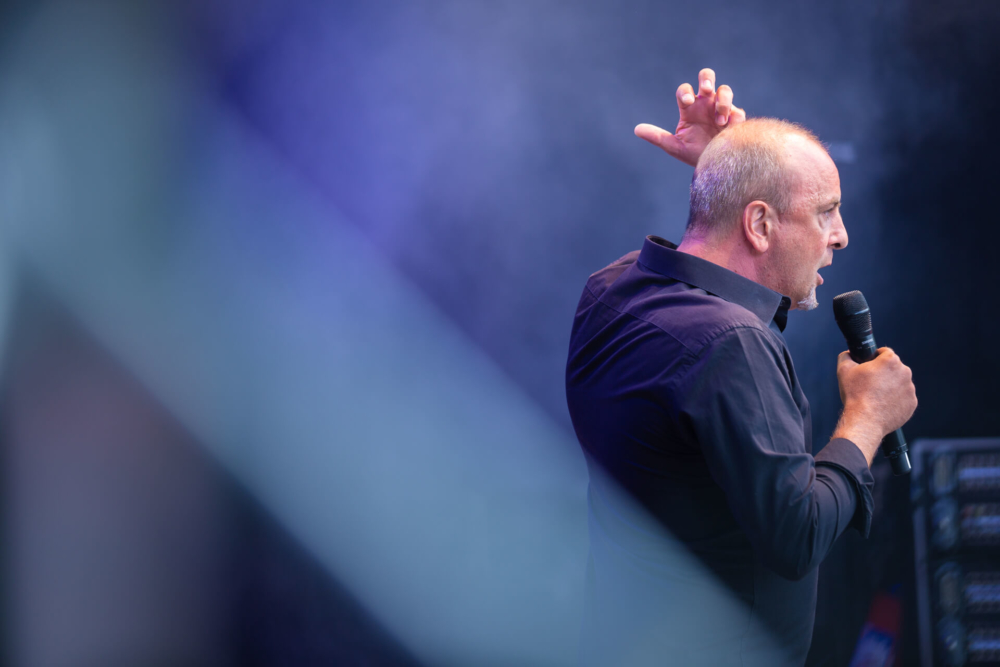 Un homme chauve se produit énergiquement sur scène avec un microphone. La scène est éclairée par des lumières bleues spectaculaires et il y a de la fumée en arrière-plan.