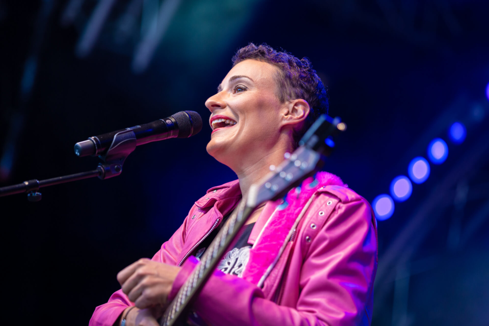 Une musicienne vêtue d'une veste rose joue sur scène avec le sourire. Elle tient un microphone et une guitare, sur fond de lumières vives.