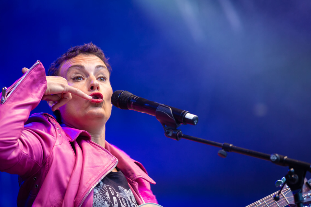 Une femme vêtue d’une veste rose chante dans un microphone sur une scène aux lumières bleues. Elle fait des gestes comme si elle était en communication téléphonique.