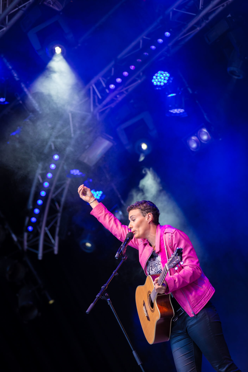 Un musicien vêtu d'une veste rose et d'un jean joue de la guitare et chante dans un micro sur scène, sous des lumières bleues.