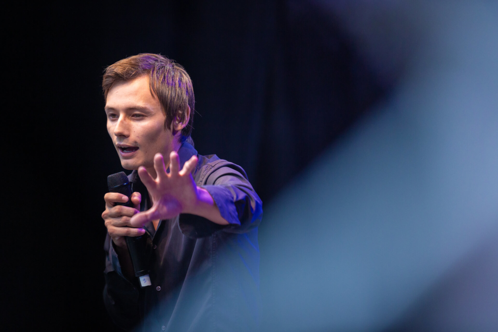 Un homme parle dans un micro sur scène. Les mouvements de ses mains sont en partie masqués par un premier plan flou. Il a l’air concentré et expressif sous les lumières vives de la scène.