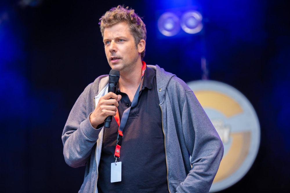 Un homme blond parle dans un microphone sur scène lors d’un événement en plein air. Il porte une veste bleue sur un sweat à capuche gris et a un air sérieux.