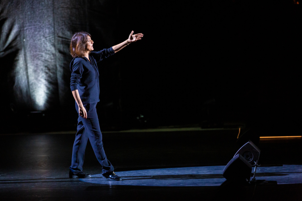 Une femme aux cheveux jusqu'aux épaules se tient sur une scène peu éclairée, étend un bras dans un mouvement théâtral, vêtue de vêtements noirs.