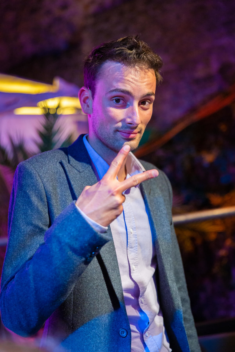 Un jeune homme surpris lors d'une fête montre un signe de paix avec sa main. Il porte une veste grise et l'arrière-plan est éclairé par des lumières colorées.