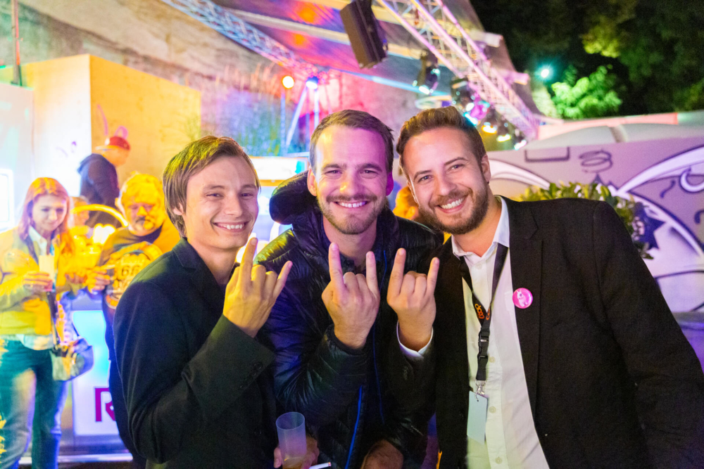Trois hommes heureux participent à un événement en plein air le soir. L'homme au milieu fait un signe de la main « rock on ». Il y a des lumières vives et d’autres personnes présentes à la fête en arrière-plan.