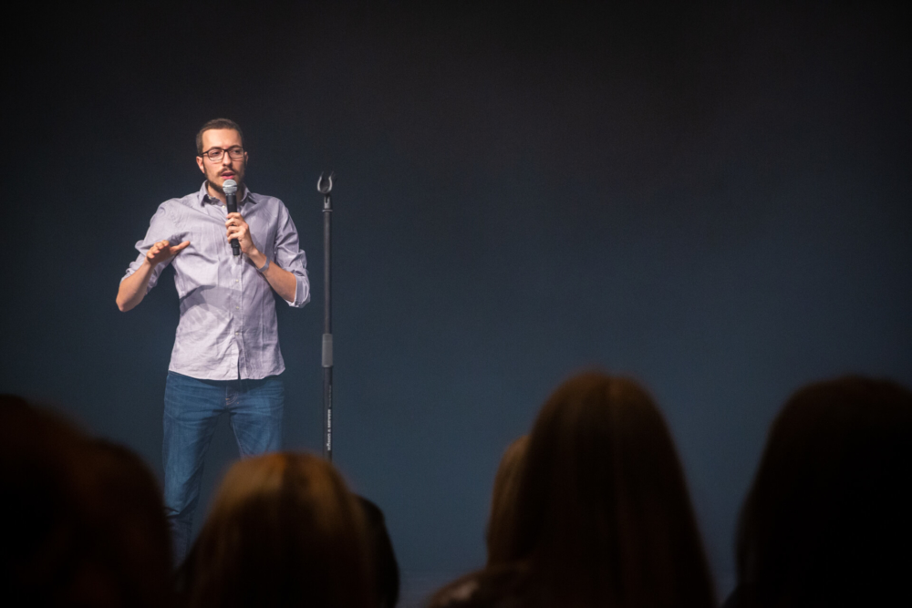Un homme, comédien, tient un micro dans une main et fait des gestes de l'autre. Il s'adresse à un auditoire dans une salle peu éclairée.