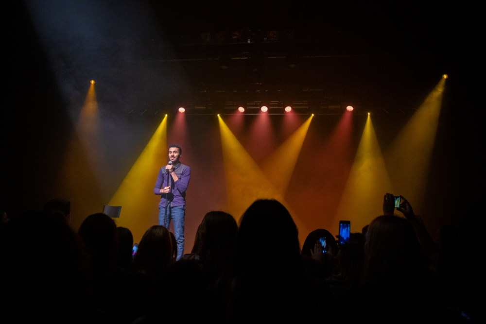 Un homme chante sur scène avec des lumières colorées qui brillent sur lui, tandis que les gens devant le filment avec leur téléphone.