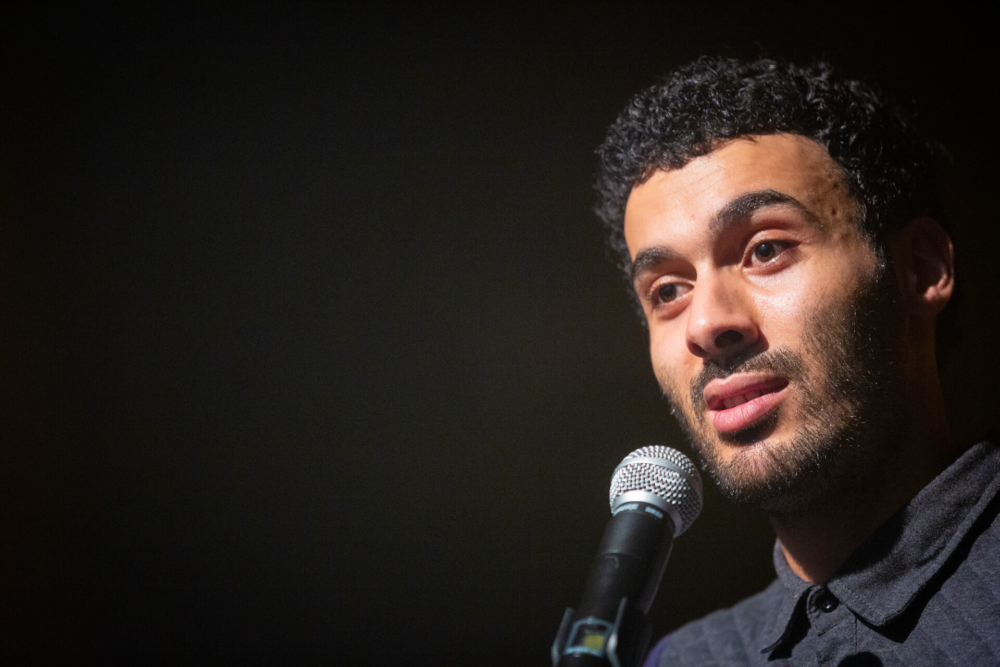 Un jeune homme parle dans un micro et attire l'attention du public par son air sérieux. Il porte une chemise grise, rendue visible par les lumières de la scène.