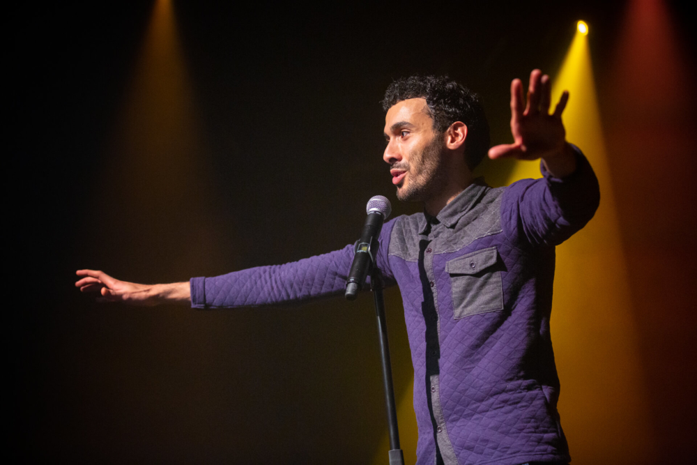 Un comédien est sur scène, faisant des gestes dramatiques avec un bras et parlant dans un microphone sous des lumières jaunes. Il porte des vêtements sombres et semble engagé auprès de son public.