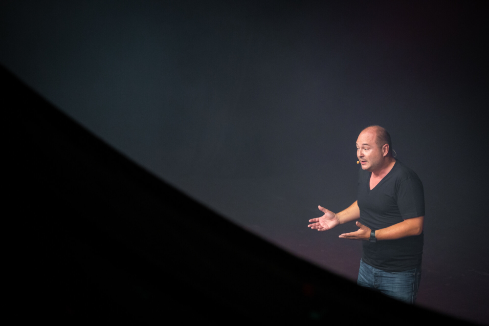 Un homme se tient sur une scène, éclairée par un projecteur, tout en faisant une présentation ou une performance. Il est encadré du côté gauche par un premier plan sombre et flou.
