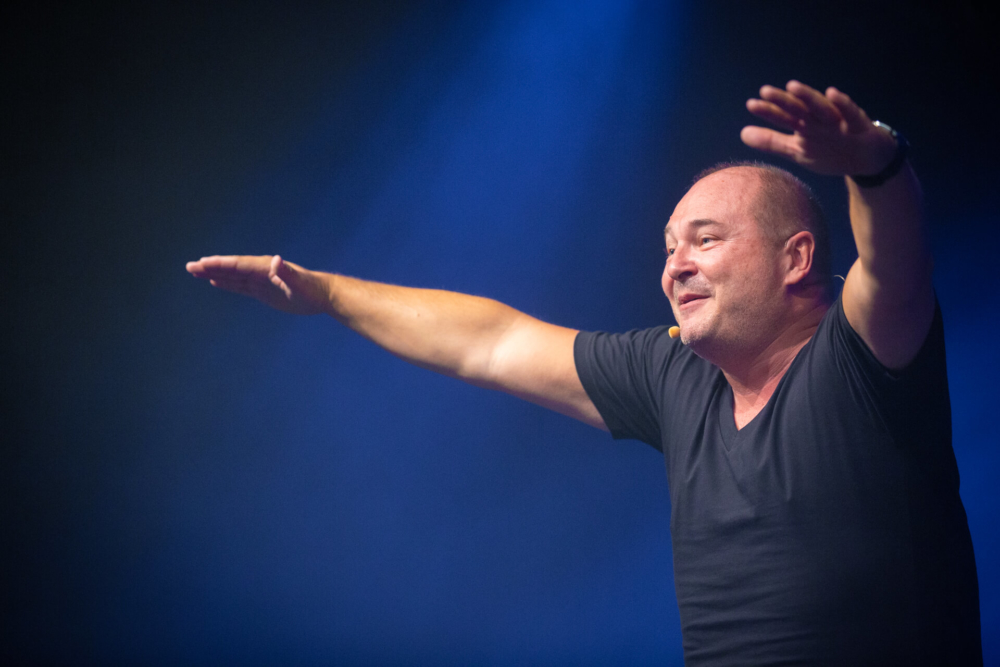 Un homme est sur scène, écartant les bras et semblant joyeux sous les lumières bleues de la scène. Il interagit avec les téléspectateurs lors d'une émission en direct.