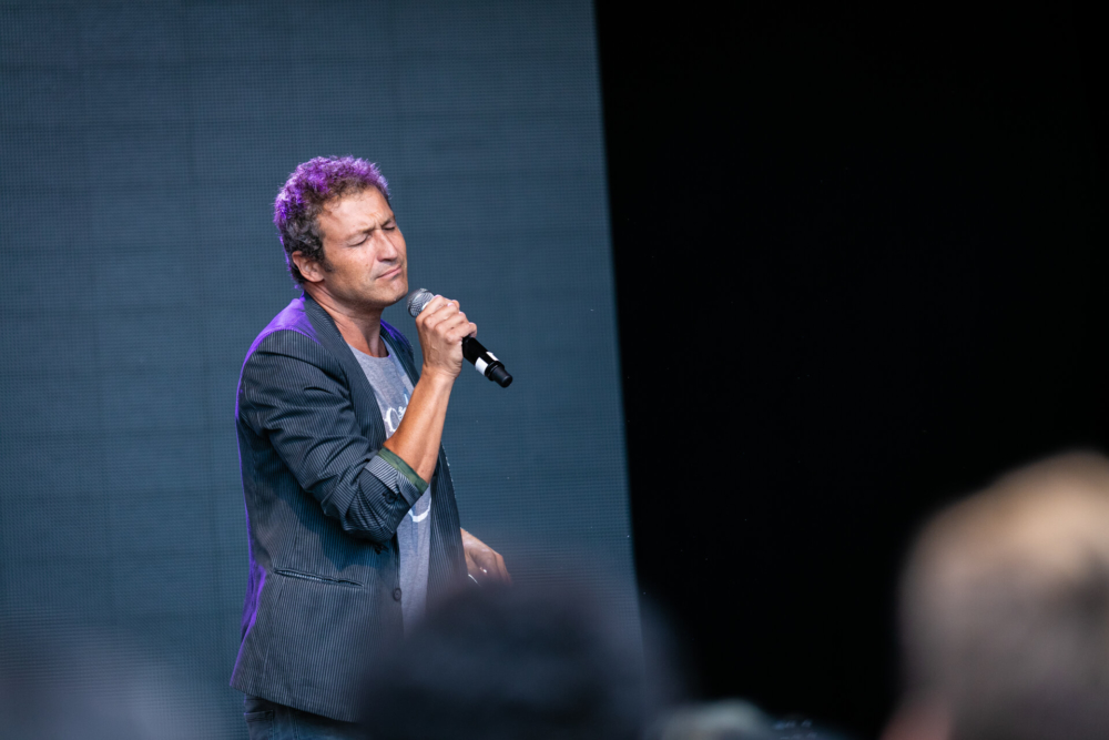 Un homme chante passionnément sur scène avec un micro. La foule devant est floue, attirant l’attention sur lui.