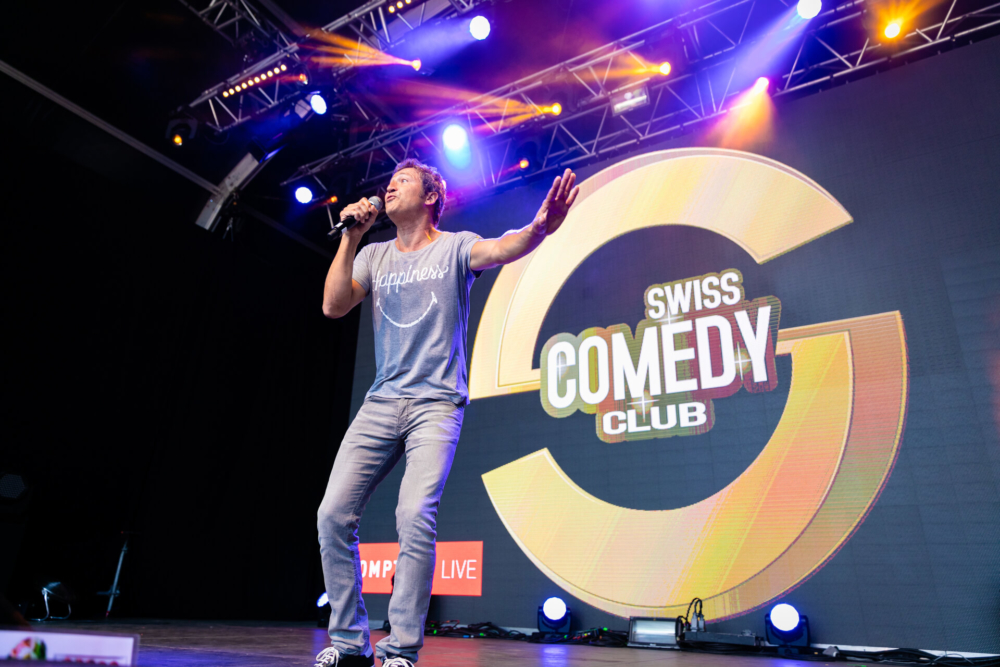 Un homme chante avec passion sur scène lors d'un événement dans un club de comédie en Suisse. La scène est éclairée par des lumières colorées et il y a un grand logo rond en arrière-plan.