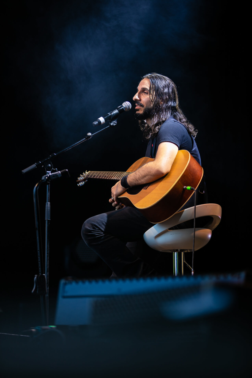 Un musicien aux cheveux longs est assis sur un tabouret, jouant d'une guitare acoustique et chantant dans un microphone sur une scène sombre avec un léger brouillard en arrière-plan.