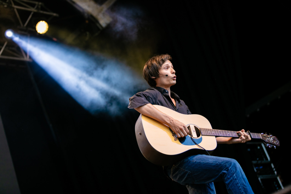 Un jeune homme joue de la guitare acoustique sur scène sous les projecteurs. Il a un regard concentré et est entouré de fumée de scène.