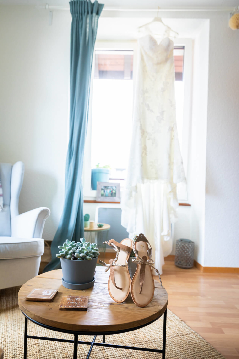 Un salon ensoleillé et confortable dispose d’une robe de mariée accrochée près de la fenêtre. Sur une table basse, il y a des sandales et une petite plante. Il existe également un livre intitulé « Plan de mariage ».