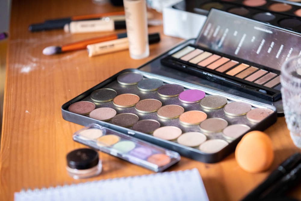 Une palette de maquillage avec des ombres à paupières de différentes couleurs, ainsi que d'autres produits de maquillage et pinceaux, se trouve sur une table en bois.