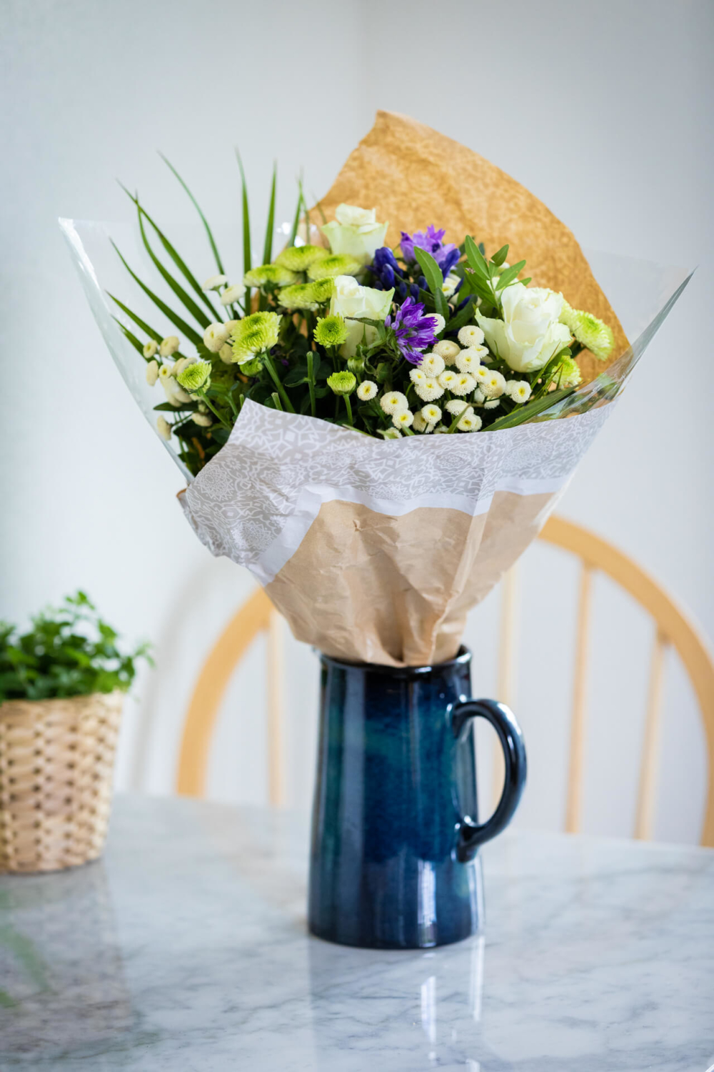 Un bouquet de fleurs colorées en papier marron et blanc se trouve dans un vase en céramique bleue. Il est sur la table de la cuisine avec un panier en osier derrière.