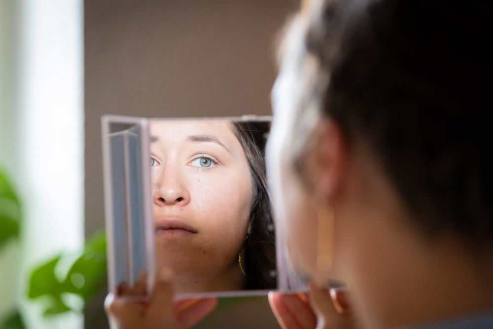 Une femme regarde profondément dans un miroir portatif, son reflet montrant un regard pensif. Ses yeux bleus sont mis en valeur par la douce lumière naturelle, soulignant son humeur réfléchie.