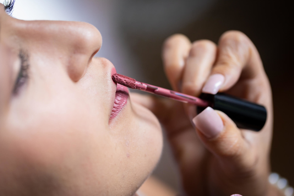 Vue rapprochée d'une femme qui se fait mettre du rouge à lèvres sur les lèvres par quelqu'un d'autre avec un pinceau à lèvres, mettant en valeur la texture et les détails de l'application du maquillage.