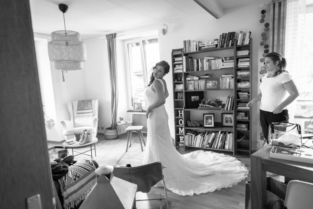 Une mariée vêtue d’une longue robe se tient debout dans un salon lumineux et lui rend son sourire. Une dame en tenue décontractée se tient à côté d’elle près d’une table en désordre.