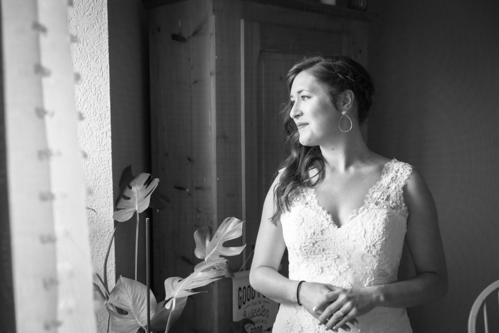 Une mariée vêtue d’une robe en dentelle regarde pensivement par la fenêtre. Elle est entourée d'une lumière douce, de belles plantes à proximité et d'un panneau indiquant « bonnes vibrations uniquement » derrière elle.