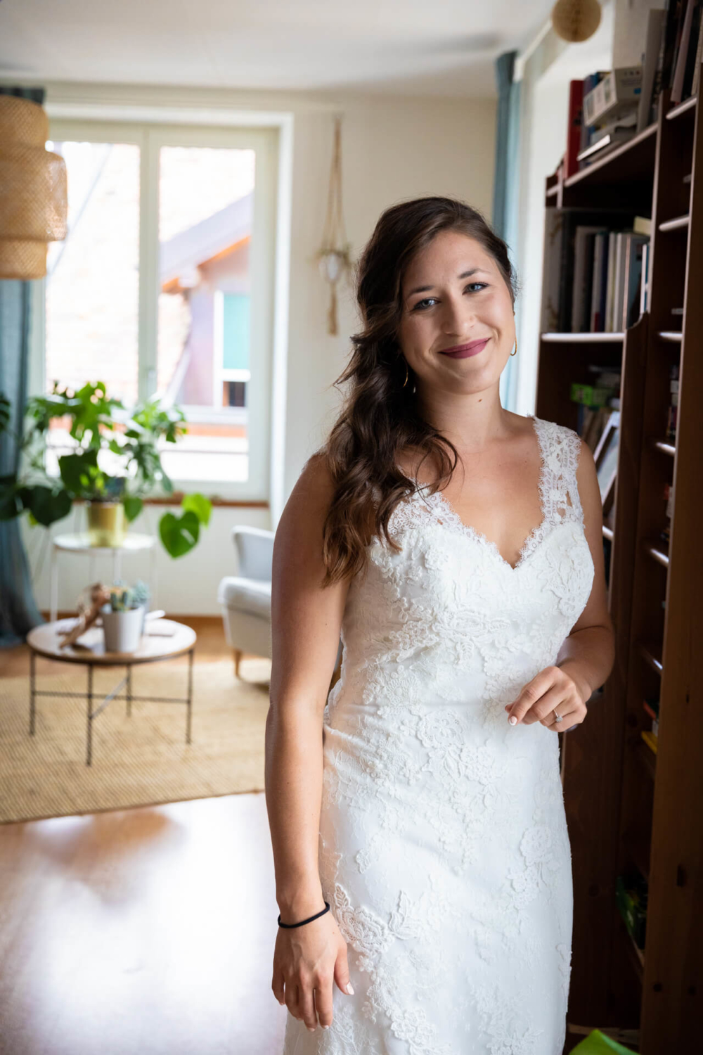 Une femme heureuse vêtue d’une robe de mariée blanche se tient dans un salon confortable avec des étagères et des décorations contemporaines.