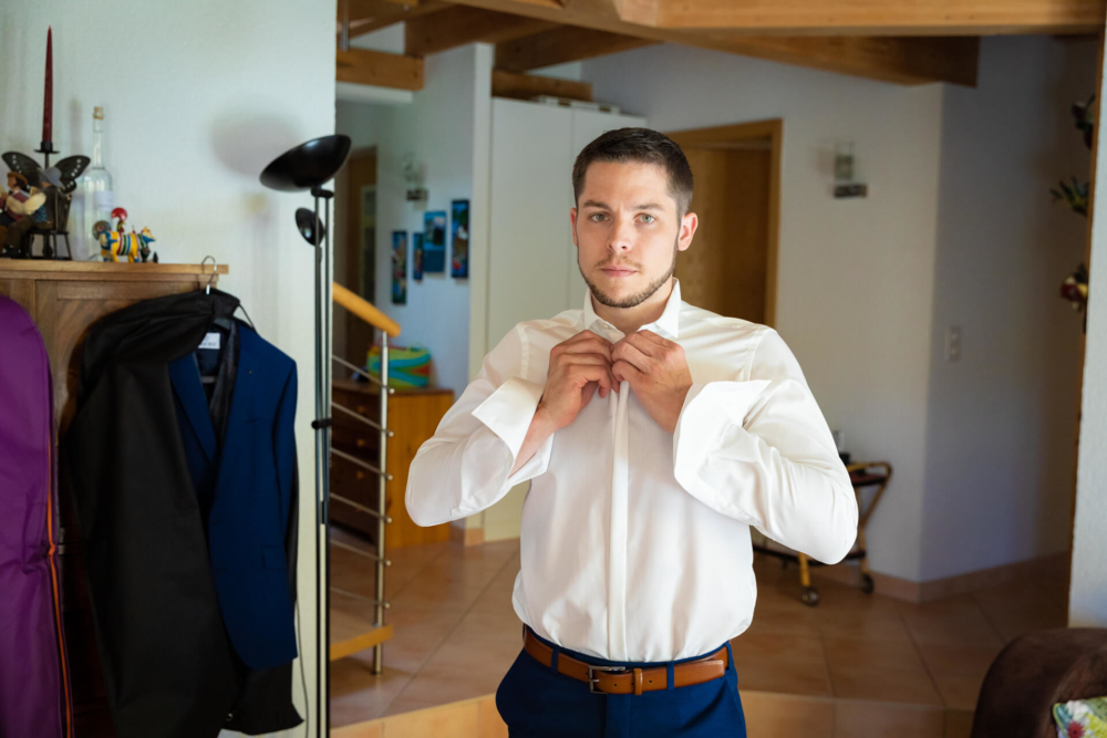 Un homme vêtu d’une chemise blanche avec une ceinture bleue attache sa cravate devant un miroir. La chambre présente une décoration en bois et un portemanteau.
