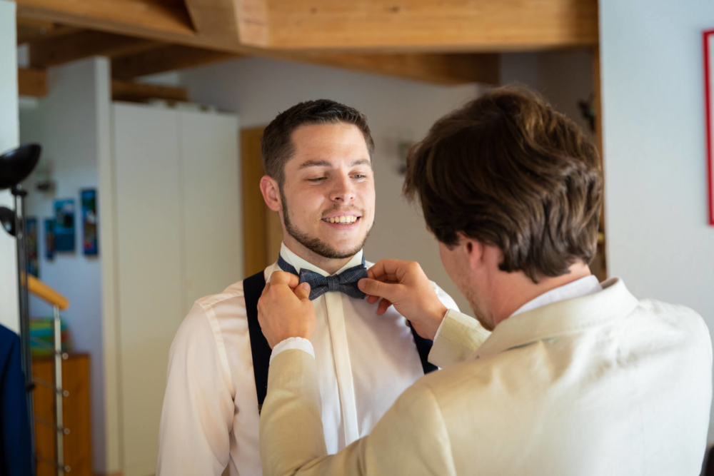 Deux hommes se trouvent dans une pièce chaleureusement éclairée avec des poutres apparentes en bois. Un homme ajuste le nœud papillon de l'autre. Ils portent tous les deux des vêtements formels, sourient et ont l'air heureux.
