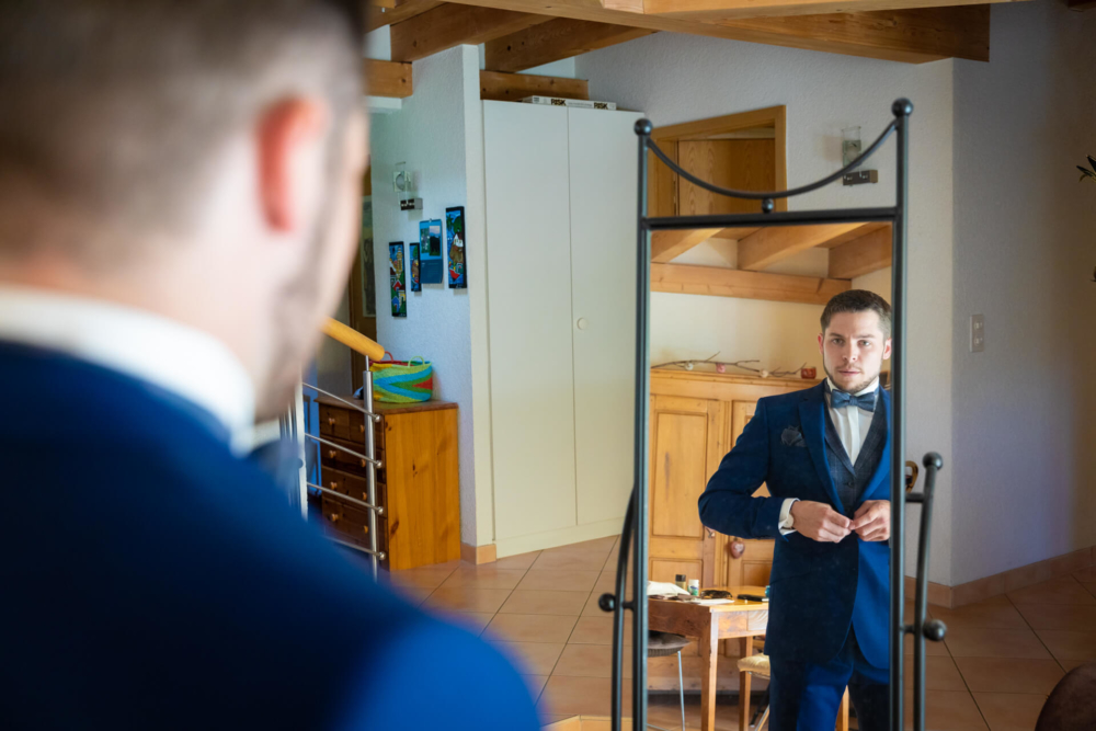 Un homme vêtu d'un costume bleu répare son bouton de manchette tout en se regardant dans un miroir. La pièce dans laquelle il se trouve est lumineuse, avec des meubles en bois et un escalier derrière lui.
