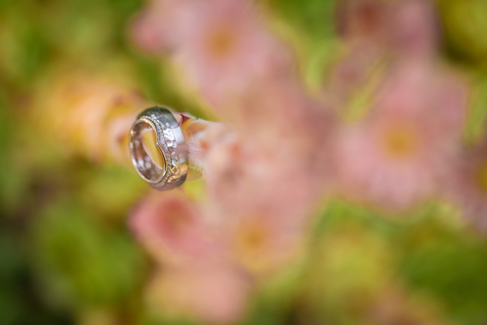 Une bague en argent détaillée avec des motifs complexes est présentée en gros plan, avec des fleurs roses floues en arrière-plan.