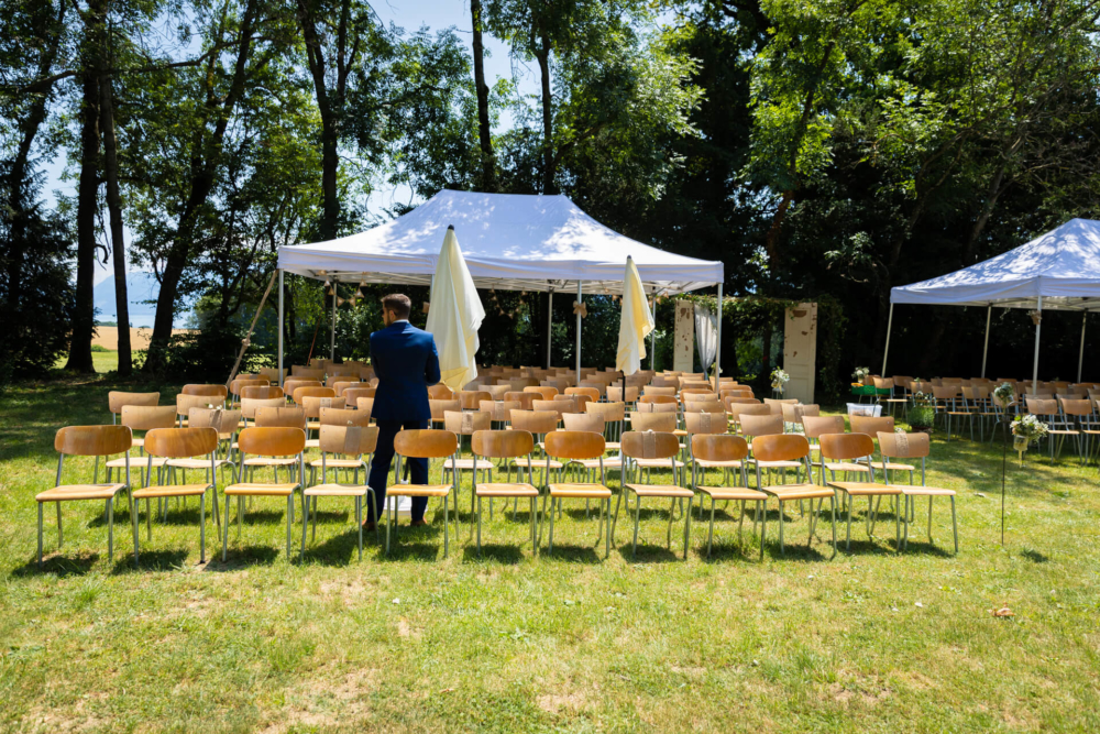 Un homme en costume se tient au bout d’une rangée de chaises marron vides disposées à l’extérieur. Ils font face à un dais de mariage blanc avec des arbres et un ciel bleu en arrière-plan.
