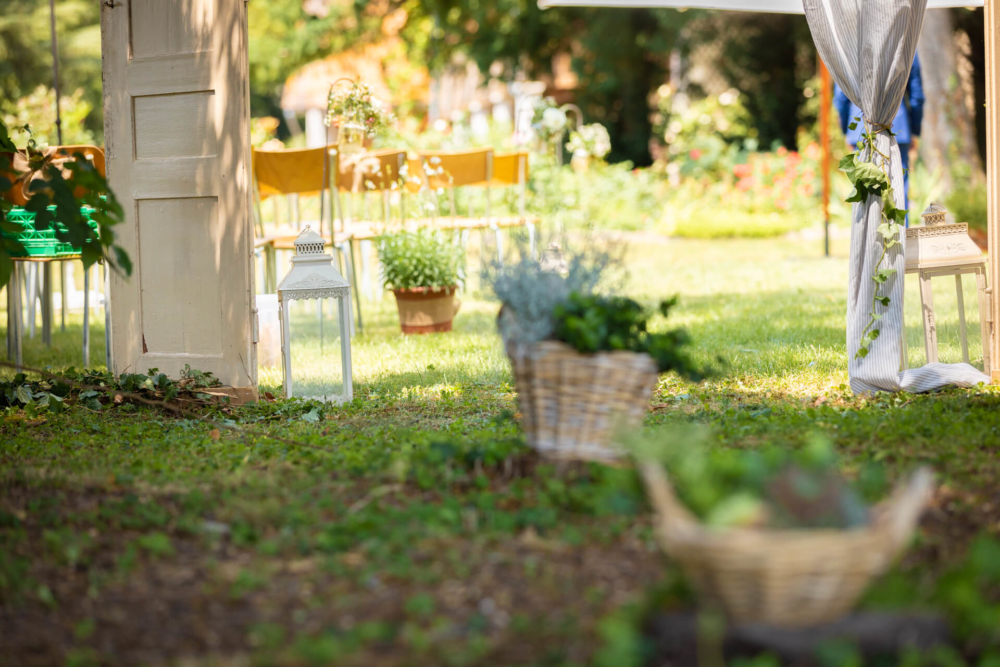 Un jardin extérieur est aménagé pour un événement avec des chaises en bois, des paniers de plantes et des lanternes, dégageant une ambiance calme et accueillante.