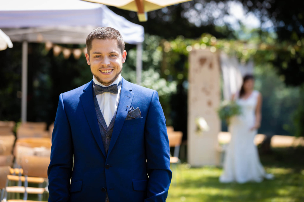 Un homme en costume bleu et nœud papillon sourit lors d'un mariage en plein air, tandis que la mariée en arrière-plan, sous un auvent ensoleillé, est légèrement floue.