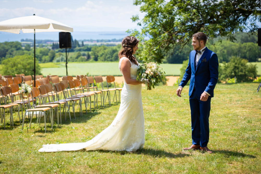 Une mariée, dans sa robe blanche fluide avec un bouquet, se tient en face du marié qui porte un costume bleu. Ils assistent à une cérémonie de mariage à l'extérieur où les chaises sont disposées en rangées. Le fond présente un magnifique paysage verdoyant.