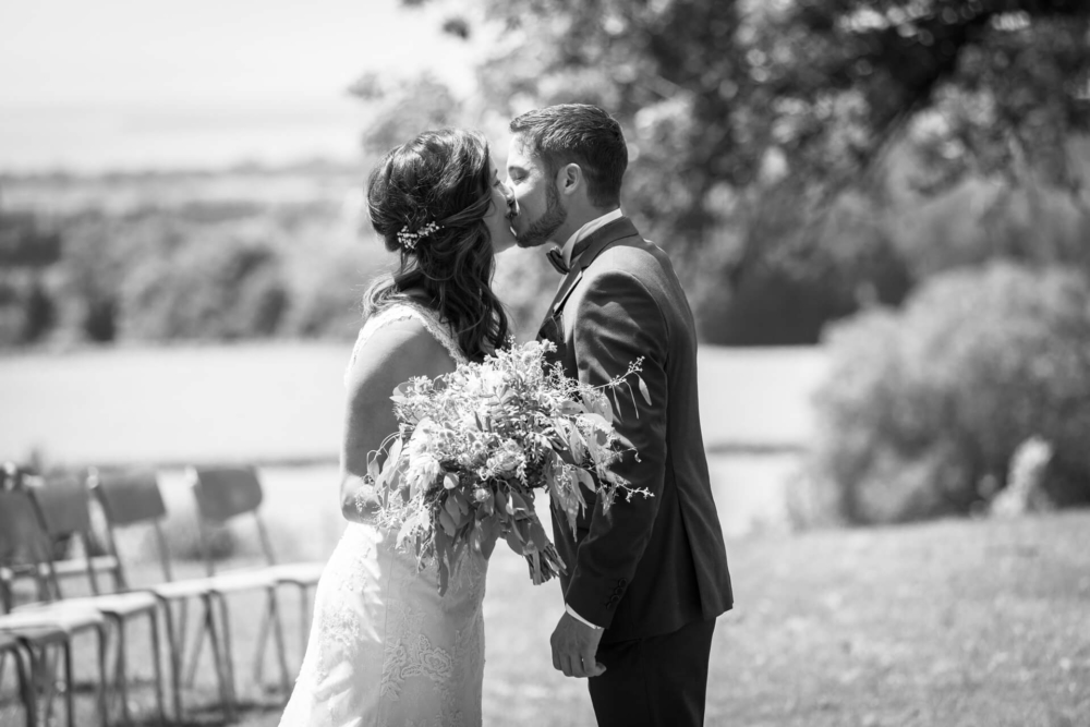 Une image en noir et blanc d’un couple nouvellement marié s’embrassant dehors avec la mariée tenant un gros bouquet. L'arrière-plan présente des paysages naturels et des chaises.