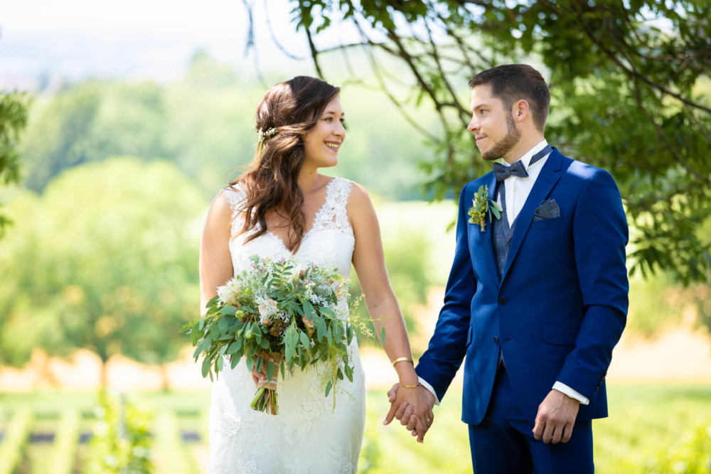 Une mariée vêtue d’une robe en dentelle blanche et un marié en costume bleu se tiennent la main sous un arbre. Ils se sourient alors que le soleil brille sur le paysage derrière eux. Le marié a également une boutonnière.